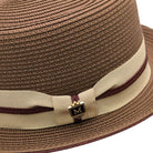 Close-up of cappuccino Montique pork pie straw hat showing woven texture, beige band, burgundy cord trim, and logo pin